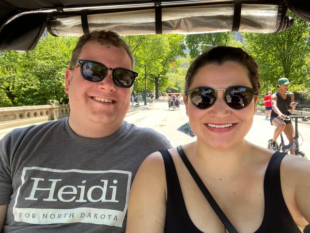 Two people smiling under a green awning on a sunny day outdoors.