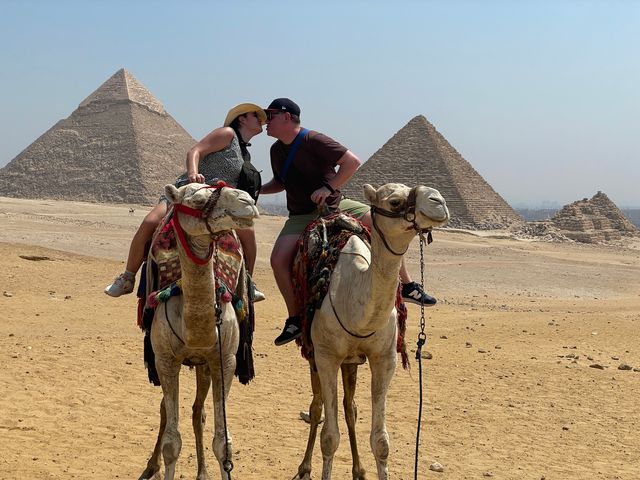 Two people on camels in front of the Egyptian pyramids in the desert.