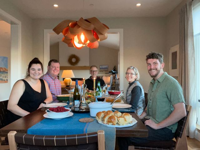Seven people gathered around a dinner table with an orange pendant lamp overhead.