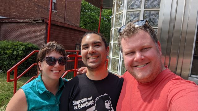 Three men posing together outdoors in front of a brick building with metal railings.
