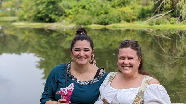 Two women smiling by a river or water body with green foliage behind them.