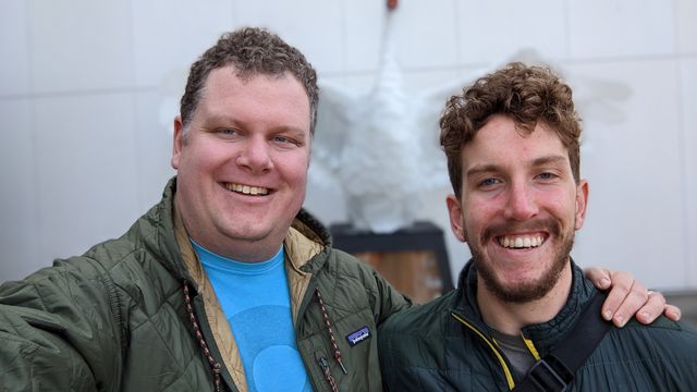 Two men smiling outdoors with steam or smoke visible in the background.