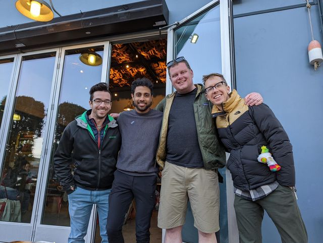 Four men smiling at the camera outside a house entrance in daylight.