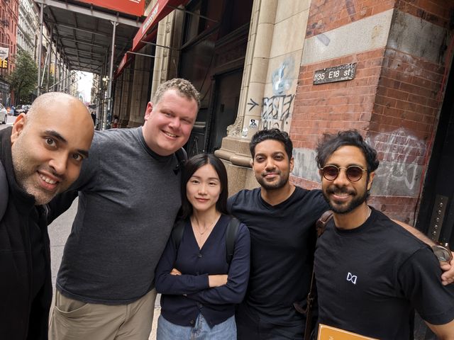 Five people taking a group selfie on a city sidewalk in front of brick buildings.