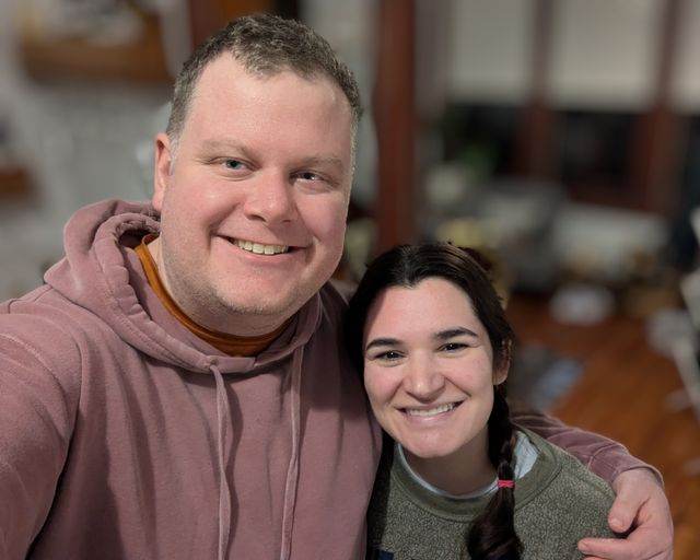 Two people smiling at the camera, one wearing a mauve hoodie and the other in a green sweater, indoors.