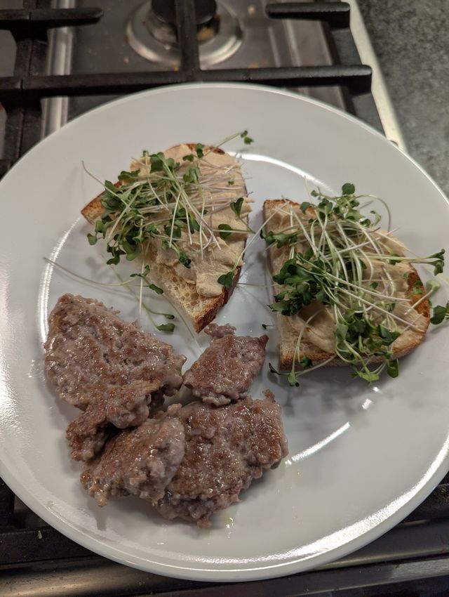 White plate with two slices of toasted sourdough topped with sprouts and hummus, accompanied by cooked breakfast sausage patties on the side.