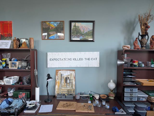 Wall display featuring framed artwork, a sign reading 'EXPECTING KILLED THE CAT,' shelves with objects, and a desk.