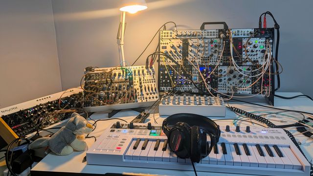 Overhead view of a white MIDI keyboard and headphones in front of several packed Eurorack cases, patch cables, a small desk lamp, and a tiny plush figure on a white desk.