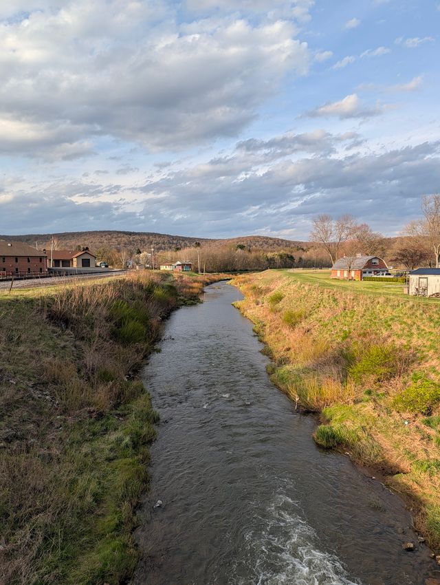 Wide stream flowing through pastoral landscape with green meadows, farm buildings, and tree-lined hills.