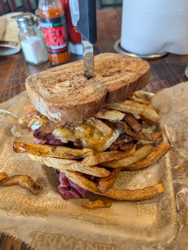 Tall stacked sandwich on Primanti-branded paper with a knife through the top, filled with crisp fries, sliced deli meat, and an egg, on a wooden table with condiments in the background.