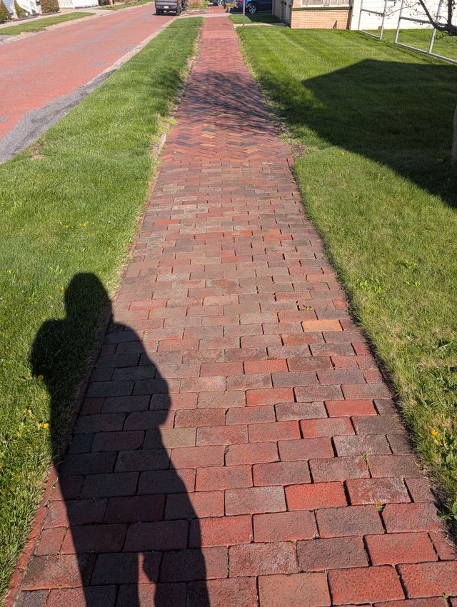 Long red-brick residential sidewalk receding into the distance in bright sun, with trimmed grass strips and a photographer's shadow in the foreground.