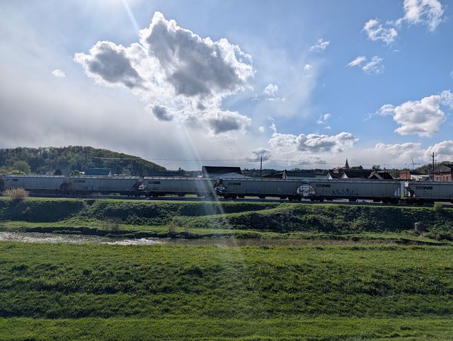 Long line of grey Norfolk Southern hopper cars on a track beside a river, green field with small yellow flowers, and a town with a church steeple under a partly cloudy sky.