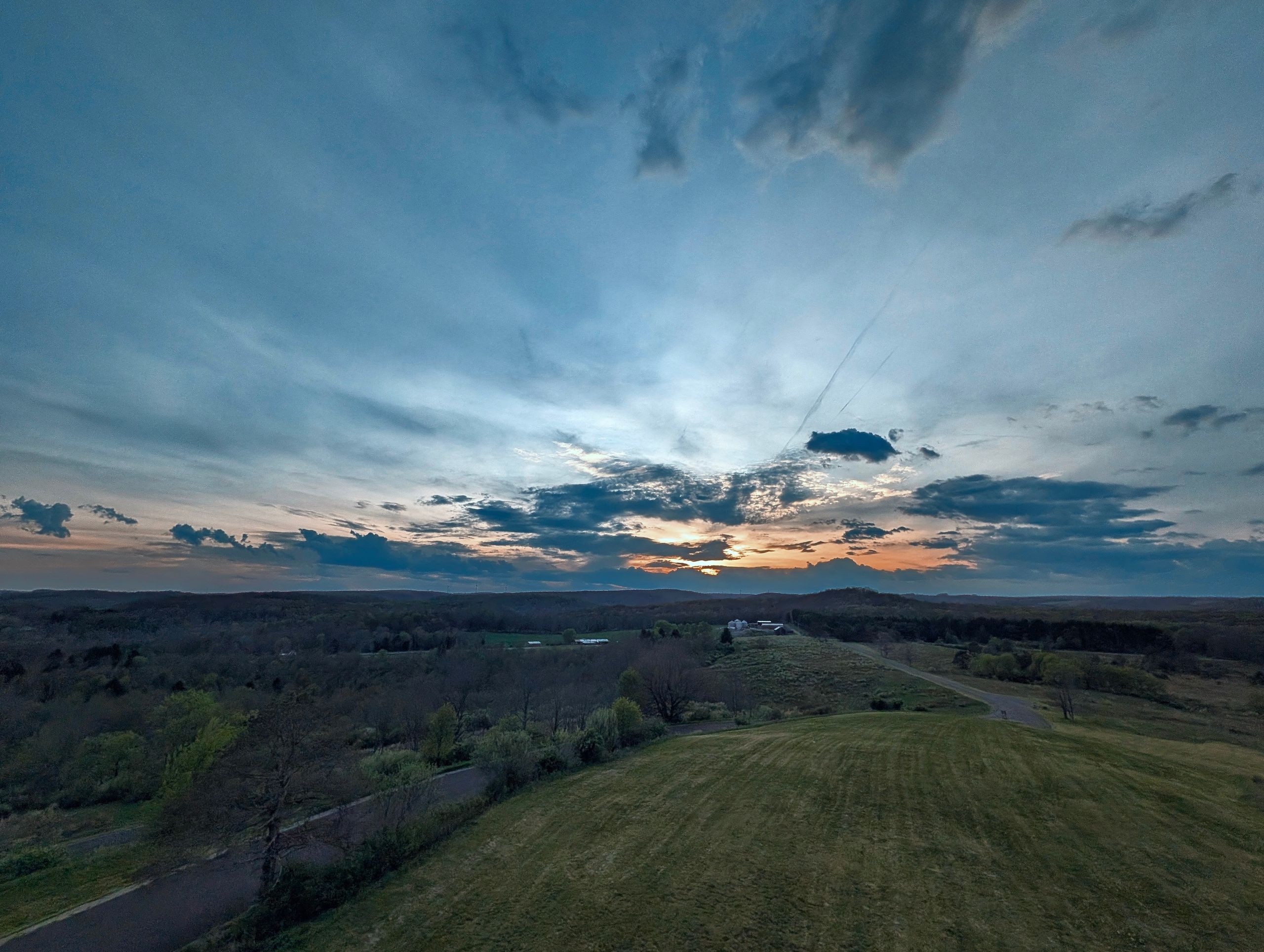 Rural scene at sunset with a deep blue sky fading to orange at the horizon, scattered clouds, a winding road, and a cluster of white farm buildings among tree-covered rolling hills.