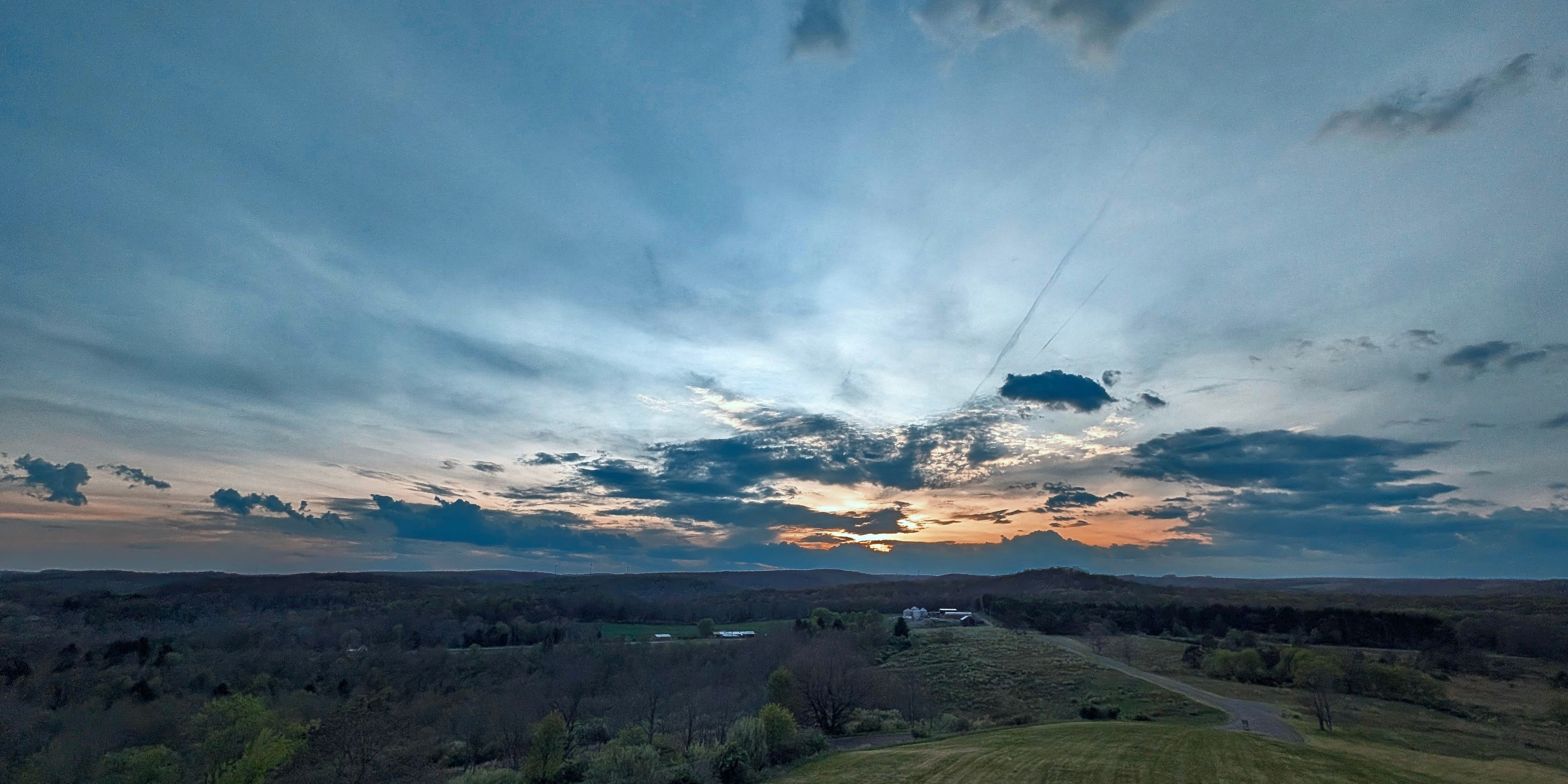 Rural scene at sunset with a deep blue sky fading to orange at the horizon, scattered clouds, a winding road, and a cluster of white farm buildings among tree-covered rolling hills.