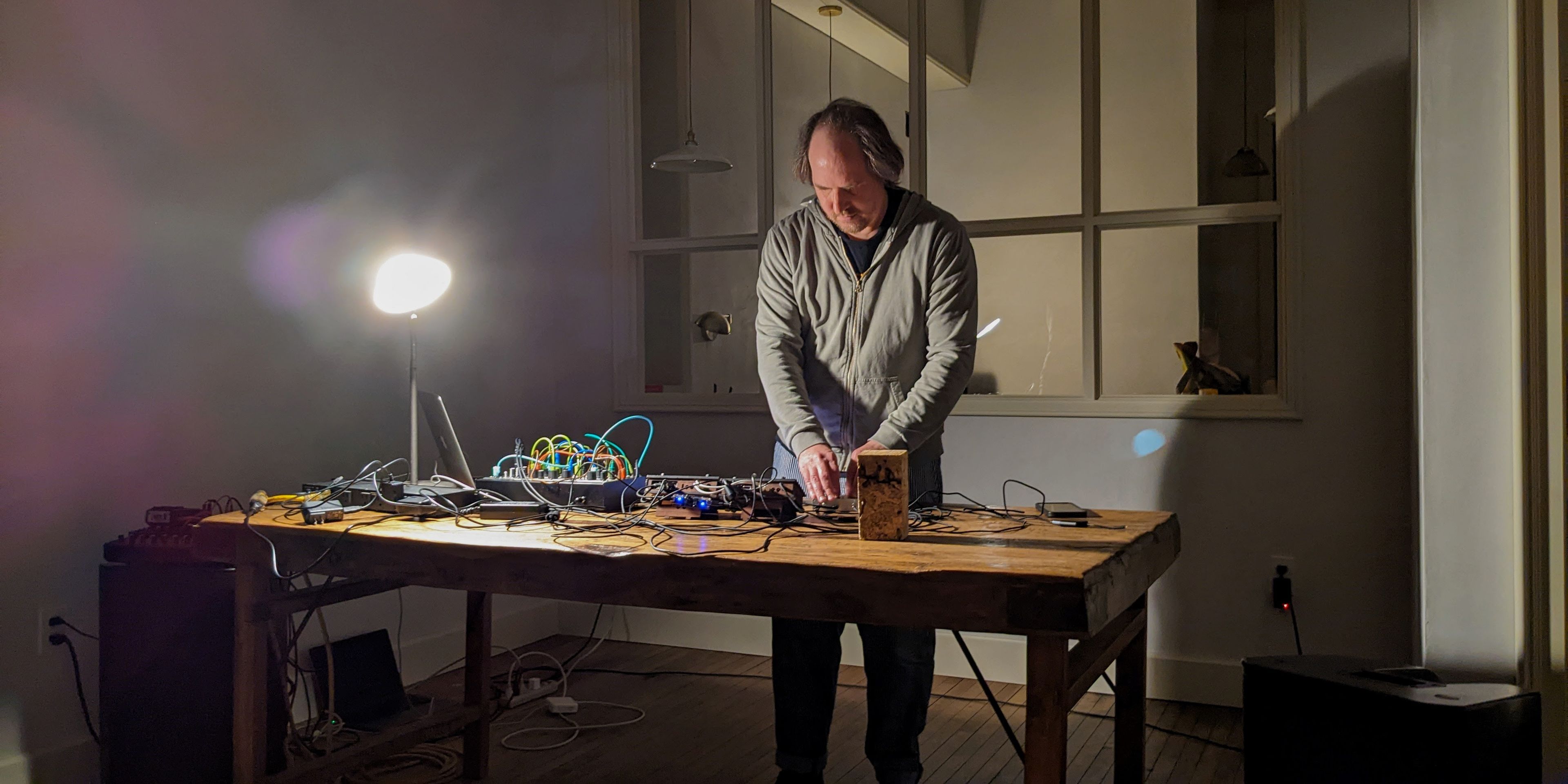A man operates electronic music equipment on a wooden table in a room.