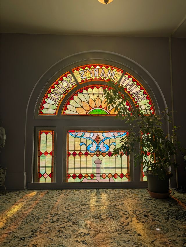 Sunlight pouring through a tall arched stained glass window labeled 'Saint Mary's School' (read in mirror image from inside), with colorful floral and geometric panels