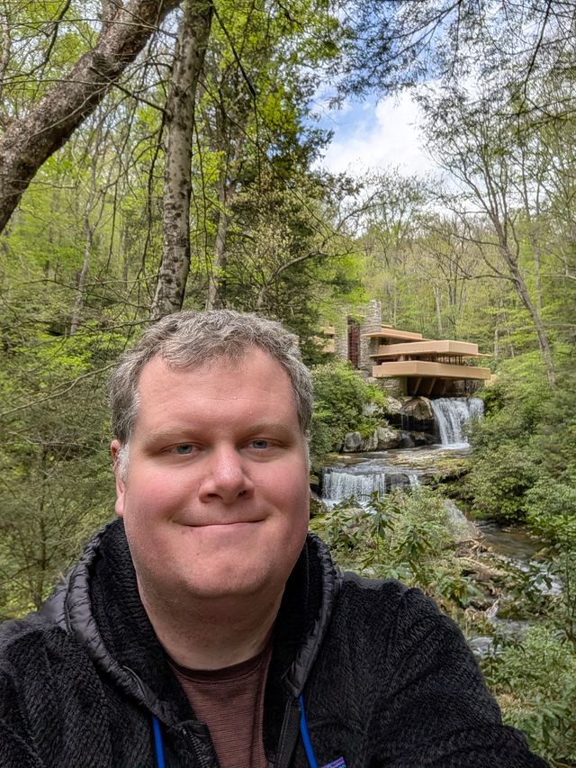 Selfie of Nat in a black fleece jacket smiling in front of Frank Lloyd Wright's Fallingwater and its waterfalls